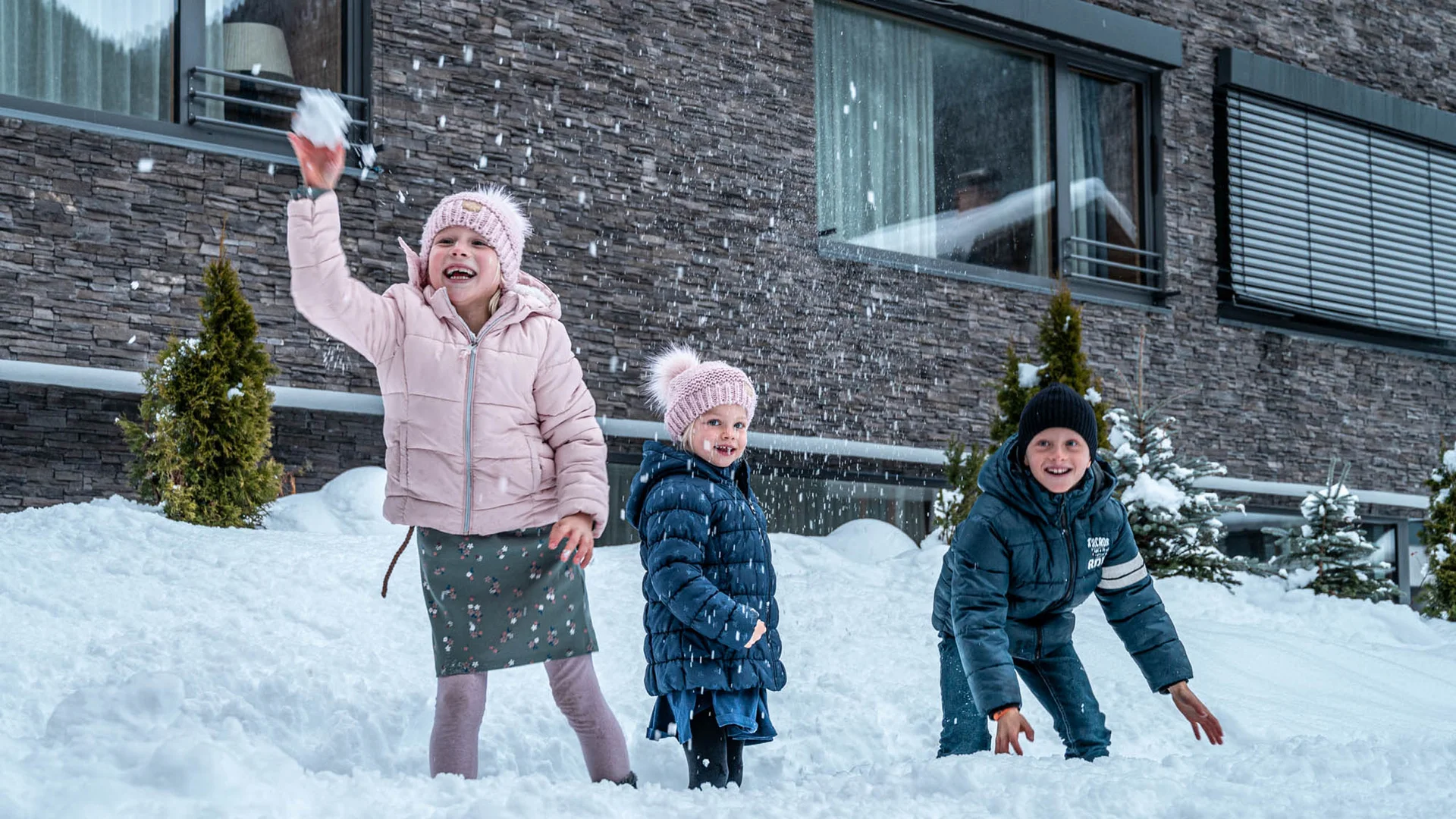 Drei Kinder spielen fröhlich im Schnee vor einem Haus